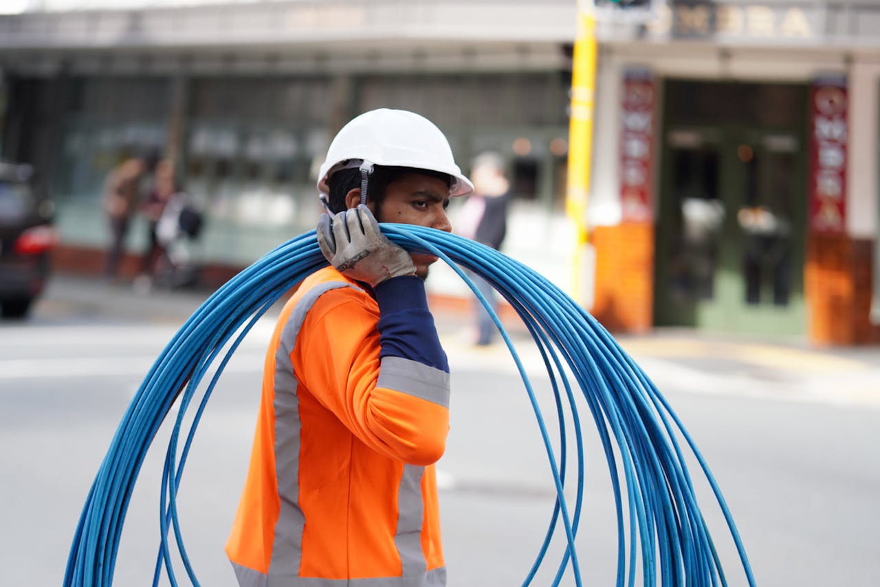 gallery-01 Construction worker in Wellington carrying blue cables, wearing safety gear on a city street.