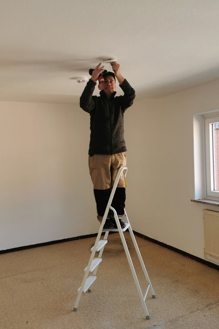 gallery-02 Adult man on ladder fixing a ceiling light in an empty room with beige walls.